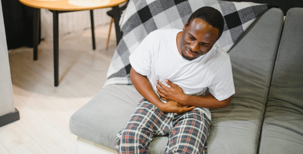 Man clutching his stomach and expressing discomfort while sitting on a sofa in a modern living room.