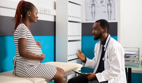 In modern medical office, young black woman discusses her pregnancy progress with friendly male physician. African american doctor seated with tablet provides details of pregnant patient checkup.