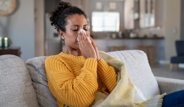 Portrait of young black woman sneezing in to tissue at home. Sick african woman wrapped in blanket sitting on sofa blowing her nose at home. Ill girl sneezing with runny nose in winter.