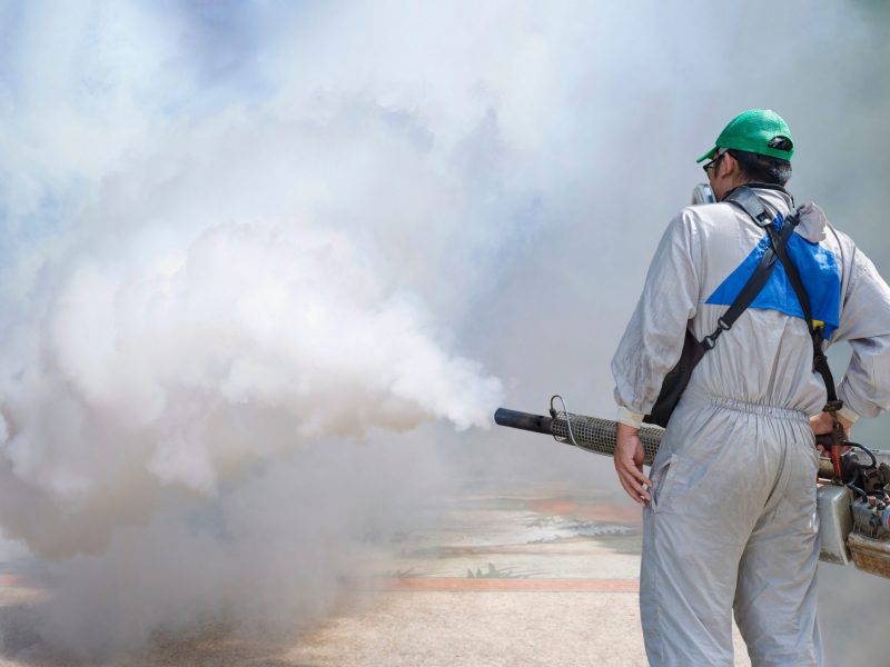 Rear view of healthcare worker in protective clothing using fogging machine spraying chemical to eliminate mosquitoes at general location in community Rear view of healthcare worker in protective clothing using fogging machine spraying chemical to eliminate mosquitoes at general location in community
